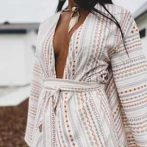 May include: A white long-sleeved dress with a deep V-neckline and a patterned design. The dress has a tie at the waist and long, flowing sleeves.