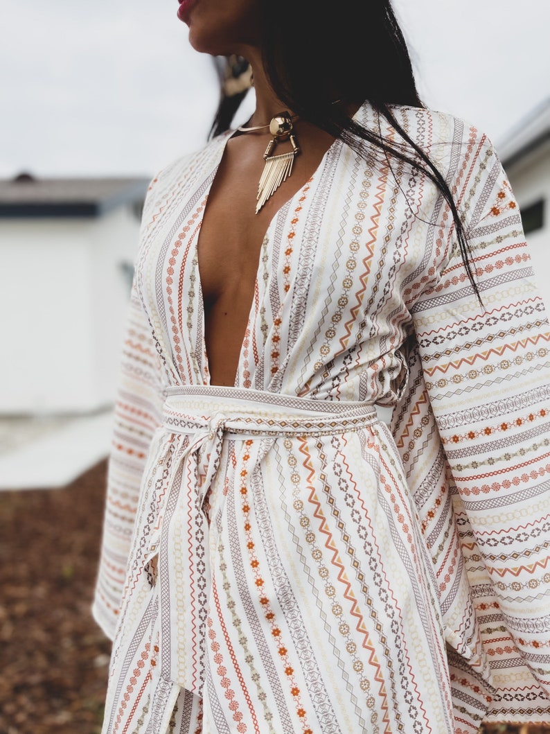 May include: A white long-sleeved dress with a deep V-neckline and a patterned design. The dress has a tie at the waist and long, flowing sleeves.