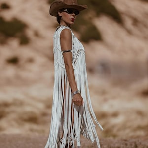 May include: A white fringed vest with silver detailing is paired with a brown cowboy hat and sunglasses. The vest features long fringe and a decorative neckline. Silver bracelets and brown boots complete the bohemian western ensemble.