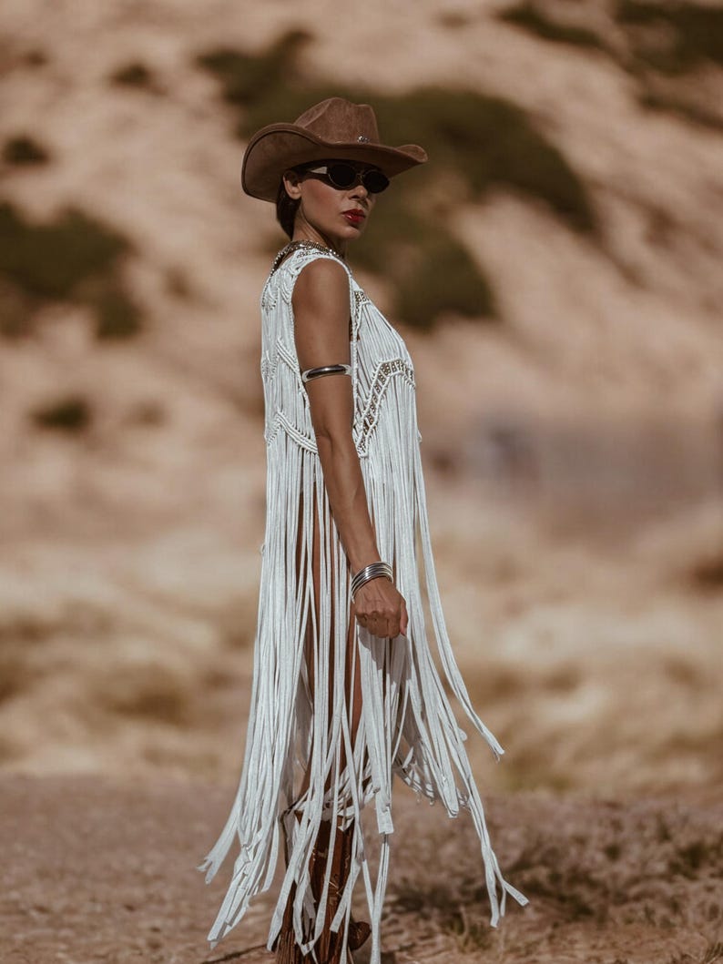 May include: A white fringed vest with silver detailing is paired with a brown cowboy hat and sunglasses. The vest features long fringe and a decorative neckline. Silver bracelets and brown boots complete the bohemian western ensemble.