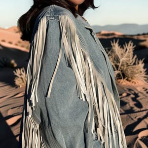 May include: A light blue denim jacket featuring long, white fringe detailing along the shoulders and sleeves. The jacket has a structured collar and a button-down front, with the fringe extending down the length of the sleeves and back panel.