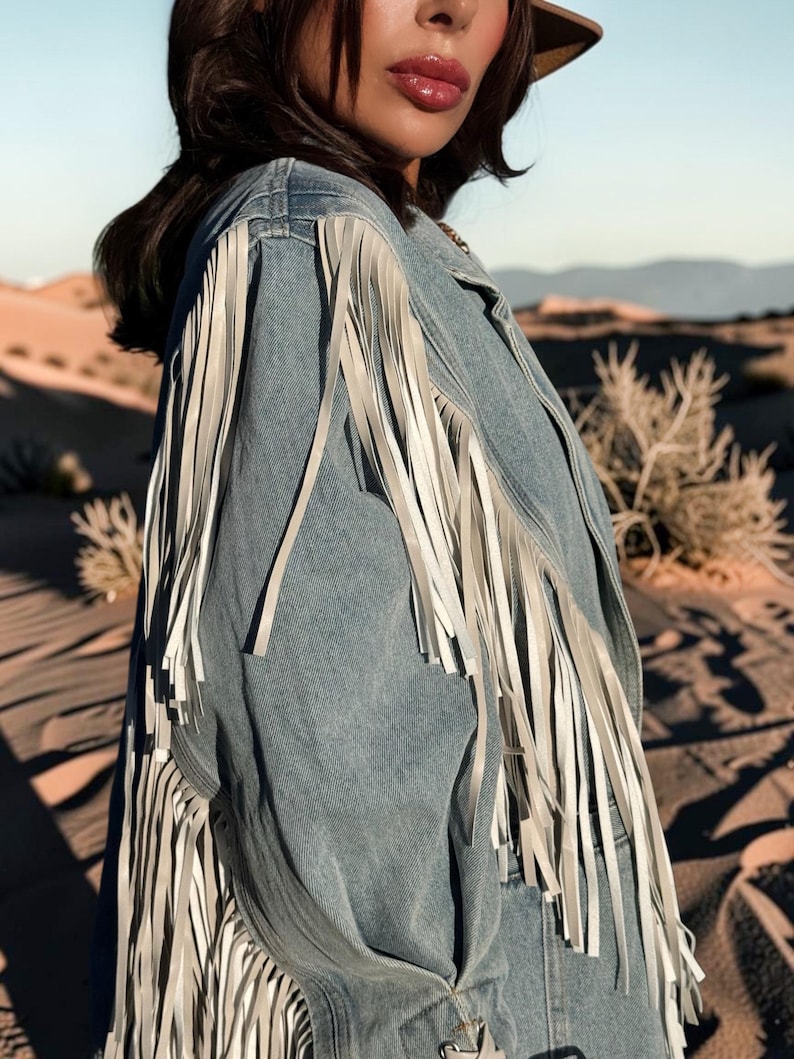 May include: A light blue denim jacket featuring long, white fringe detailing along the shoulders and sleeves. The jacket has a structured collar and a button-down front, with the fringe extending down the length of the sleeves and back panel.