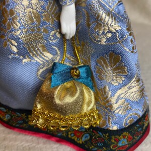 May include: A porcelain doll wearing a blue and gold patterned dress, holding a small gold and teal purse. The dress has a detailed floral pattern and a decorative trim with red accents. The doll's hand is visible.