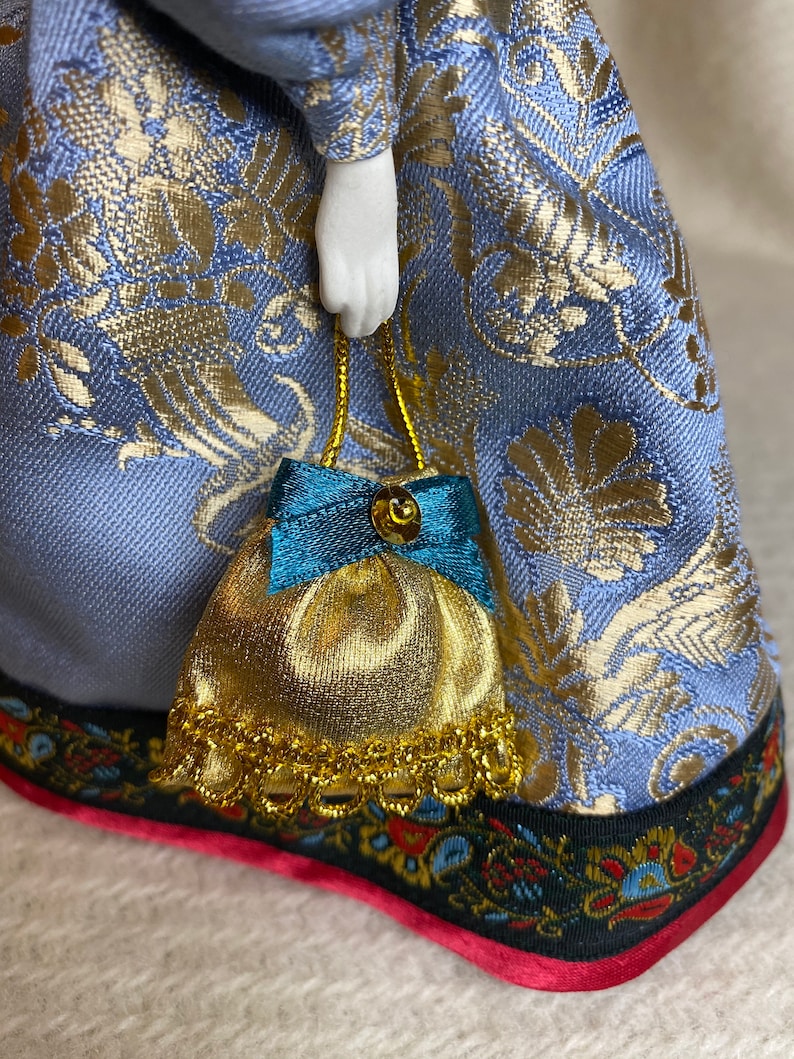 May include: A porcelain doll wearing a blue and gold patterned dress, holding a small gold and teal purse. The dress has a detailed floral pattern and a decorative trim with red accents. The doll's hand is visible.