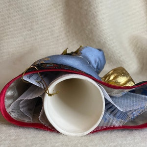 May include: A white ceramic bell with a blue and gold fabric design. The bell has a red trim and a gold string for hanging. The bell is decorated with a blue and gold fabric design.