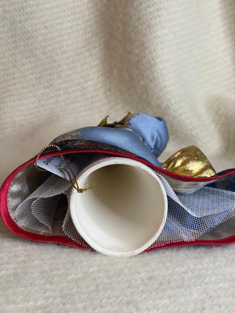 May include: A white ceramic bell with a blue and gold fabric design. The bell has a red trim and a gold string for hanging. The bell is decorated with a blue and gold fabric design.
