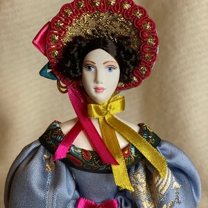 May include: A porcelain doll with a detailed face, wearing a blue dress with gold embroidery and puffed sleeves. The doll has a decorative hat with red and gold accents, and pink and gold ribbons. The doll's hair is dark and curly.