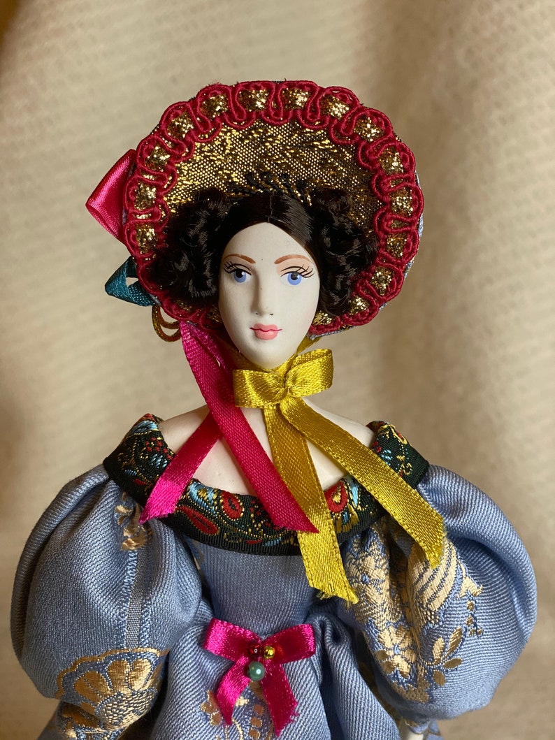 May include: A porcelain doll with a detailed face, wearing a blue dress with gold embroidery and puffed sleeves. The doll has a decorative hat with red and gold accents, and pink and gold ribbons. The doll's hair is dark and curly.