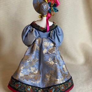 May include: A handmade doll wearing a light blue dress with gold floral patterns. The doll has a matching hat adorned with colorful ribbons and a decorative floral accent. The dress features a detailed trim with floral and paisley designs.