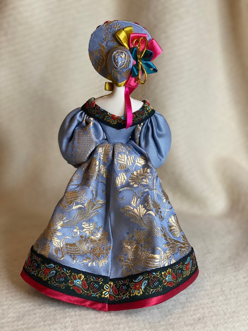 May include: A handmade doll wearing a light blue dress with gold floral patterns. The doll has a matching hat adorned with colorful ribbons and a decorative floral accent. The dress features a detailed trim with floral and paisley designs.