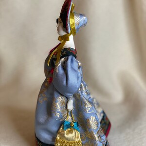 May include: A porcelain doll in a traditional Russian costume. The doll wears a light blue dress with gold floral patterns, a matching head covering, and a red and gold headdress. A small gold purse with a blue bow hangs from her hand.