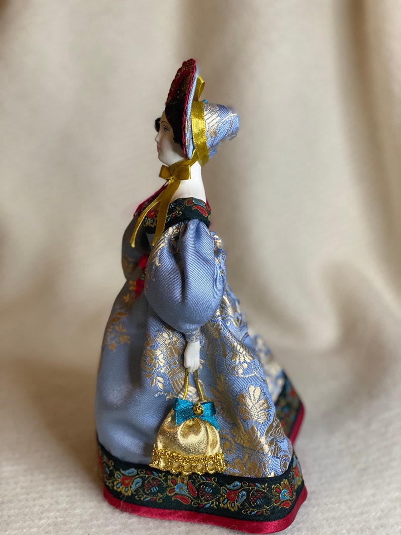May include: A porcelain doll in a traditional Russian costume. The doll wears a light blue dress with gold floral patterns, a matching head covering, and a red and gold headdress. A small gold purse with a blue bow hangs from her hand.