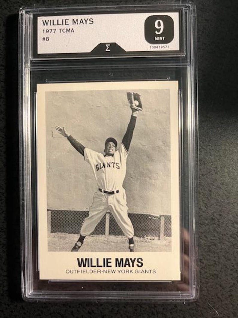 Op de afbeelding: Zwart-wit honkbalkaart van Willie Mays, 1977 TCMA, beoordeeld met Mint 9. De kaart toont Mays in een Giants-uniform, een bal vangend. Tekst bevat "WILLIE MAYS" en "OUTFIELDER-NEW YORK GIANTS".