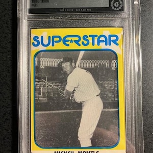 Op de afbeelding: Een vintage Mickey Mantle honkbalkaart in een beschermende houder. De kaart toont een zwart-wit afbeelding van Mantle in zijn honkbaluniform, met het woord "SUPERSTAR" in blauw op een gele achtergrond. De kaart is beoordeeld met "5 EX" door Golden Grading.