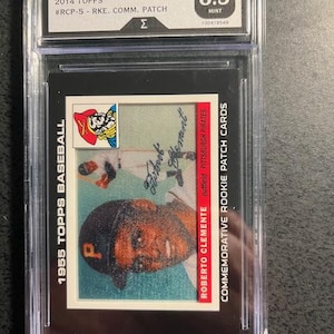 May include: A 1955 Topps baseball card featuring Roberto Clemente, graded 8.5, encased in a protective holder. The card has a portrait of Clemente in a Pittsburgh Pirates cap and the text "Roberto Clemente Commemorative Rookie Patch Cards."