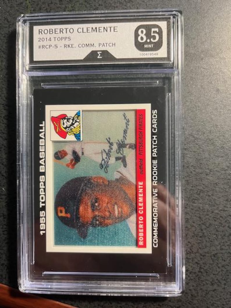 May include: A 1955 Topps baseball card featuring Roberto Clemente, graded 8.5, encased in a protective holder. The card has a portrait of Clemente in a Pittsburgh Pirates cap and the text "Roberto Clemente Commemorative Rookie Patch Cards."