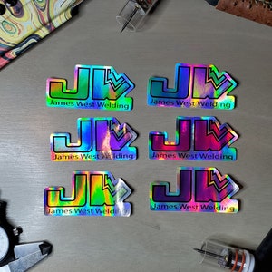 May include: Six iridescent holographic stickers with the text "James West Welding" and a stylized "JW" logo. The stickers are rainbow colored with a variety of color combinations.
