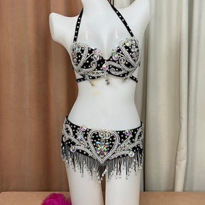 May include: A black and silver belly dance costume with a halter top and a hip belt. The costume is heavily embellished with rhinestones and dangling beads. The bra and belt feature intricate designs and a heart-shaped motif. A mannequin displays the costume.