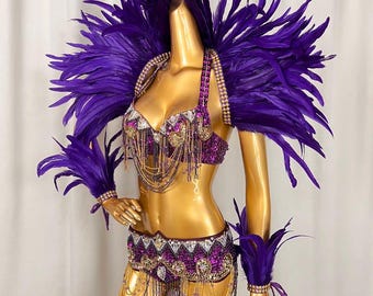 May include: A vibrant purple costume with extensive feather embellishments. The outfit features a jeweled bra, a matching belt with dangling beads, and feathered shoulder pieces and wrist cuffs. The mannequin is gold.