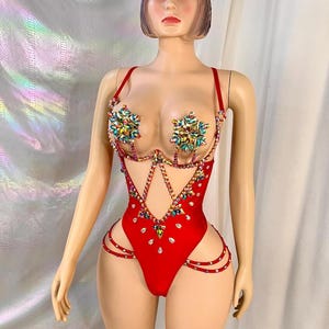 May include: A red bodysuit with rhinestone embellishments. The costume features a halter-style top with floral-shaped rhinestone accents and cutouts along the waist and hips. The bodysuit is displayed on a mannequin.