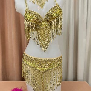 May include: A gold sequined belly dance costume set on a white mannequin. The set includes a bra, belt, and choker, all adorned with gold sequins and beaded fringe. The bra and belt have intricate designs, and the choker has a matching fringe.