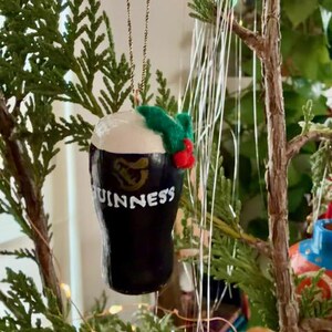 Pode incluir: Um enfeite de Natal em forma de caneca de Guinness. O enfeite preto e branco tem o logotipo e o texto da Guinness. &Eacute; decorado com um raminho de azevinho verde e vermelho e pendurado por um fio dourado. O enfeite &eacute; exibido em uma &aacute;rvore de Natal.