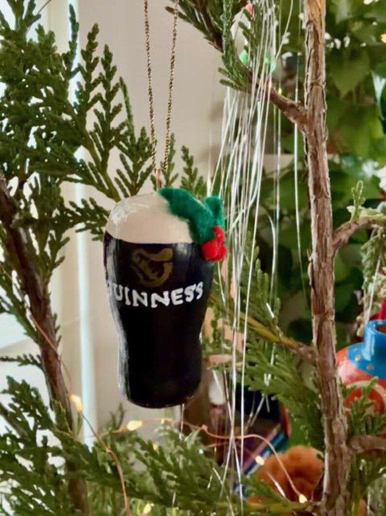 Pode incluir: Um enfeite de Natal em forma de caneca de Guinness. O enfeite preto e branco tem o logotipo e o texto da Guinness. &Eacute; decorado com um raminho de azevinho verde e vermelho e pendurado por um fio dourado. O enfeite &eacute; exibido em uma &aacute;rvore de Natal.