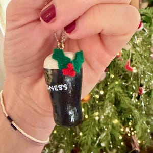 Pode incluir: Um colar com pingente de cor escura com uma miniatura de Guinness com espuma branca e detalhes de azevinho verde e vermelho. A palavra "Guinness" &eacute; vis&iacute;vel na caneca. O colar &eacute; preso por uma corrente dourada.