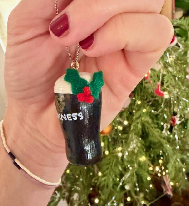 Pode incluir: Um colar com pingente de cor escura com uma miniatura de Guinness com espuma branca e detalhes de azevinho verde e vermelho. A palavra "Guinness" &eacute; vis&iacute;vel na caneca. O colar &eacute; preso por uma corrente dourada.