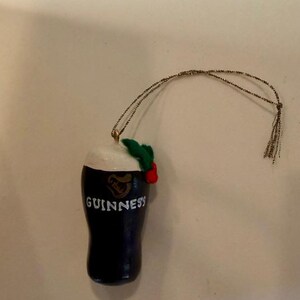 Pode incluir: Enfeite de Natal em forma de caneca Guinness azul escuro com espuma branca, o logotipo Guinness e a palavra "Guinness" em branco. O enfeite &eacute; decorado com folhas verdes e bagas vermelhas e tem um fio dourado para pendurar.