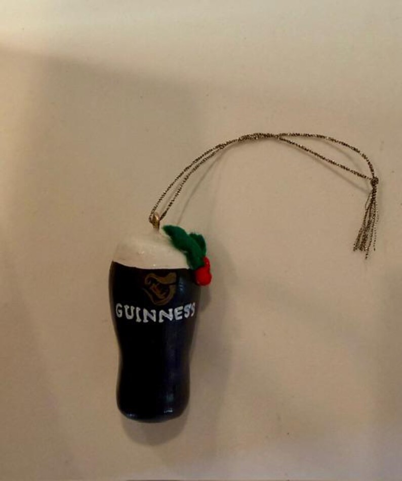 Pode incluir: Enfeite de Natal em forma de caneca Guinness azul escuro com espuma branca, o logotipo Guinness e a palavra "Guinness" em branco. O enfeite &eacute; decorado com folhas verdes e bagas vermelhas e tem um fio dourado para pendurar.