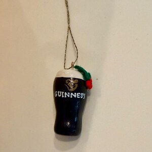 Pode incluir: Um enfeite de caneca Guinness azul marinho escuro com uma espuma branca no topo. A palavra "GUINNESS" &eacute; impressa em branco na frente. Um raminho verde com uma baga vermelha &eacute; preso no topo. O enfeite pende de um fio dourado.