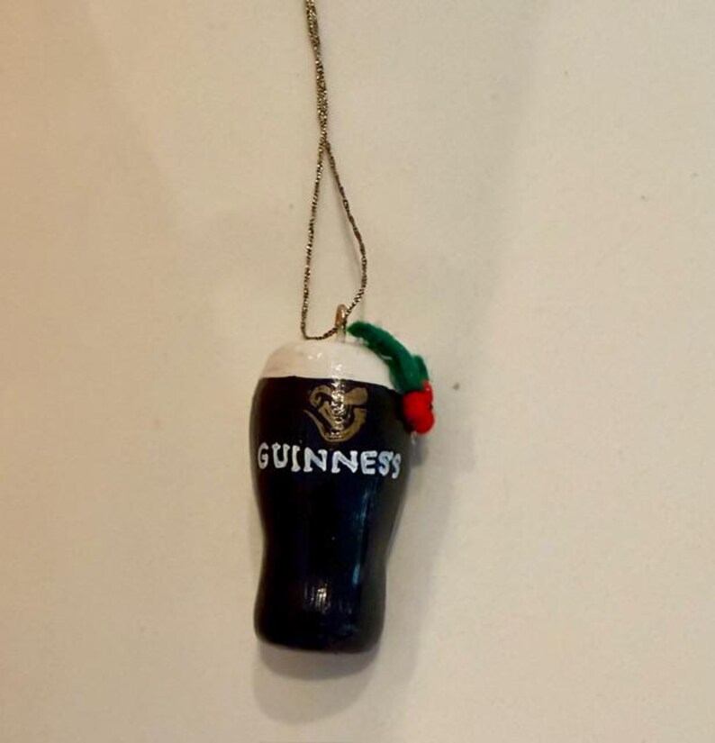 Pode incluir: Um enfeite de caneca Guinness azul marinho escuro com uma espuma branca no topo. A palavra "GUINNESS" &eacute; impressa em branco na frente. Um raminho verde com uma baga vermelha &eacute; preso no topo. O enfeite pende de um fio dourado.