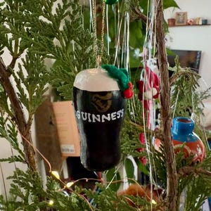 Pode incluir: Um enfeite de Natal em forma de caneca de Guinness. O enfeite &eacute; preto com espuma branca e o logotipo da Guinness. &Eacute; decorado com um pequeno ramo verde e bagas vermelhas, suspenso por uma corrente dourada.