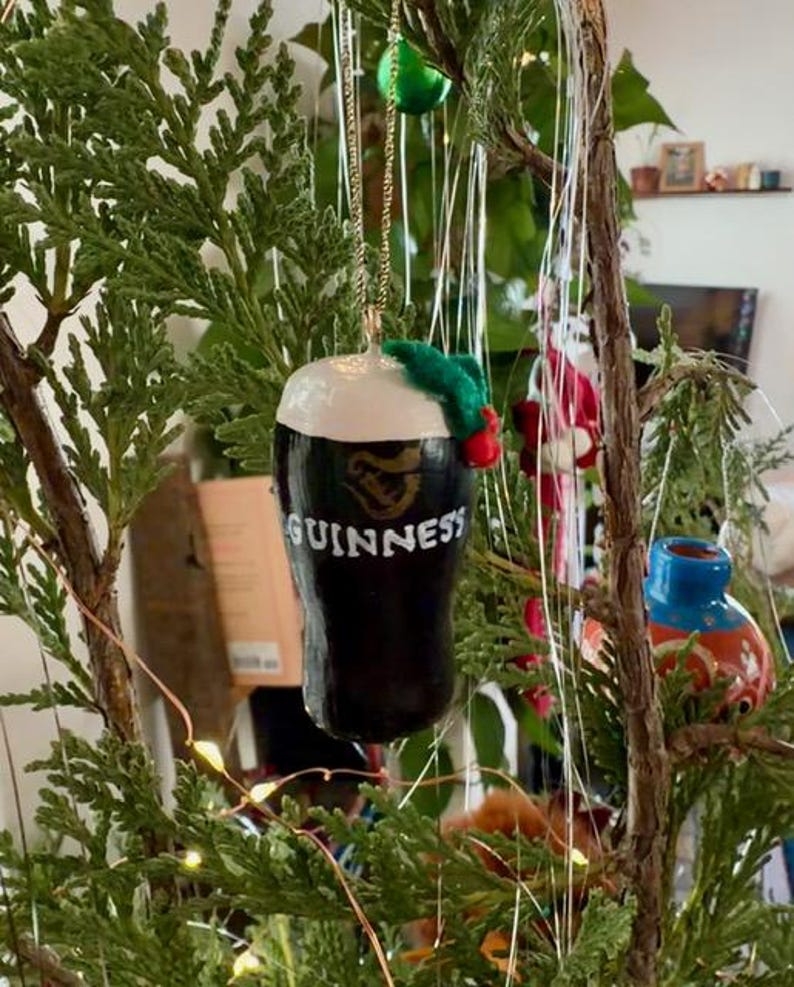 Pode incluir: Um enfeite de Natal em forma de caneca de Guinness. O enfeite &eacute; preto com espuma branca e o logotipo da Guinness. &Eacute; decorado com um pequeno ramo verde e bagas vermelhas, suspenso por uma corrente dourada.