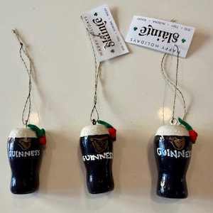 Pode incluir: Tr&ecirc;s enfeites de Natal em forma de caneca de Guinness. Cada um &eacute; azul escuro com uma parte superior de espuma branca, o logotipo da Guinness e a palavra "Guinness" em branco. Cada um tem uma folha verde e uma baga vermelha.