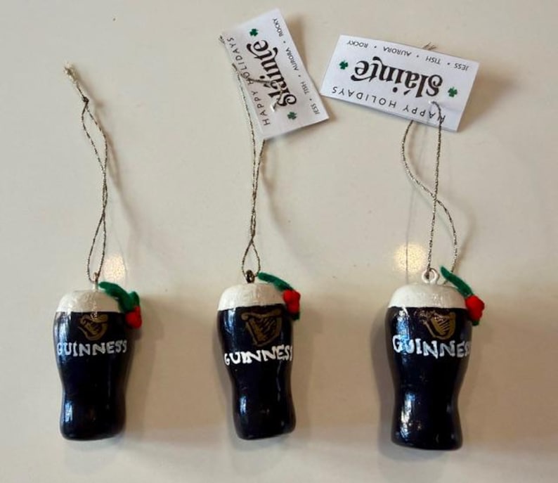 Pode incluir: Tr&ecirc;s enfeites de Natal em forma de caneca de Guinness. Cada um &eacute; azul escuro com uma parte superior de espuma branca, o logotipo da Guinness e a palavra "Guinness" em branco. Cada um tem uma folha verde e uma baga vermelha.