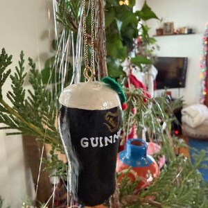 Pode incluir: Um enfeite de Natal em forma de copo de Guinness. O enfeite preto tem uma cabe&ccedil;a branca e o logotipo da Guinness. Ele pende de uma corrente dourada e &eacute; decorado com um pequeno galho verde.