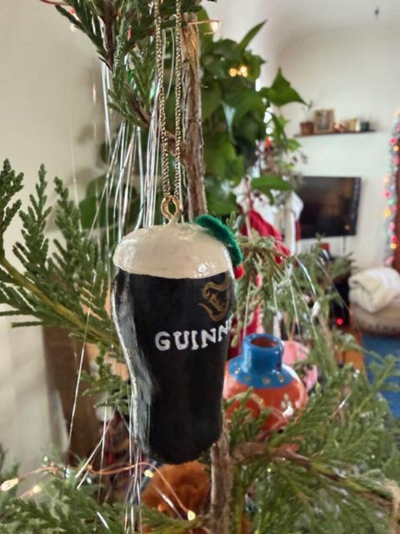 Pode incluir: Um enfeite de Natal em forma de copo de Guinness. O enfeite preto tem uma cabe&ccedil;a branca e o logotipo da Guinness. Ele pende de uma corrente dourada e &eacute; decorado com um pequeno galho verde.