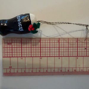 Pode incluir: Um enfeite em miniatura de um copo de Guinness. O copo preto tem a palavra "Guinness" em branco, com uma harpa dourada. O enfeite tem uma cabe&ccedil;a branca e &eacute; decorado com folhas verdes e bagas vermelhas. O enfeite tem cerca de 5 cm de comprimento.