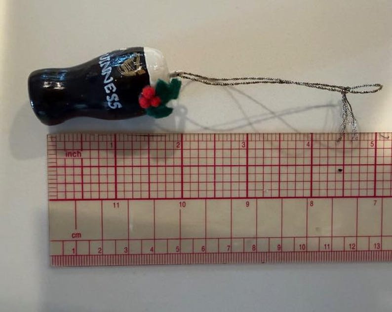 Pode incluir: Um enfeite em miniatura de um copo de Guinness. O copo preto tem a palavra "Guinness" em branco, com uma harpa dourada. O enfeite tem uma cabe&ccedil;a branca e &eacute; decorado com folhas verdes e bagas vermelhas. O enfeite tem cerca de 5 cm de comprimento.