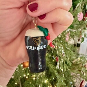 Pode incluir: Um enfeite de Natal de copo de cerveja Guinness preto e branco com uma espuma branca. O enfeite tem a palavra "Guinness" em letras brancas. Um raminho de azevinho verde e vermelho est&aacute; preso no topo.