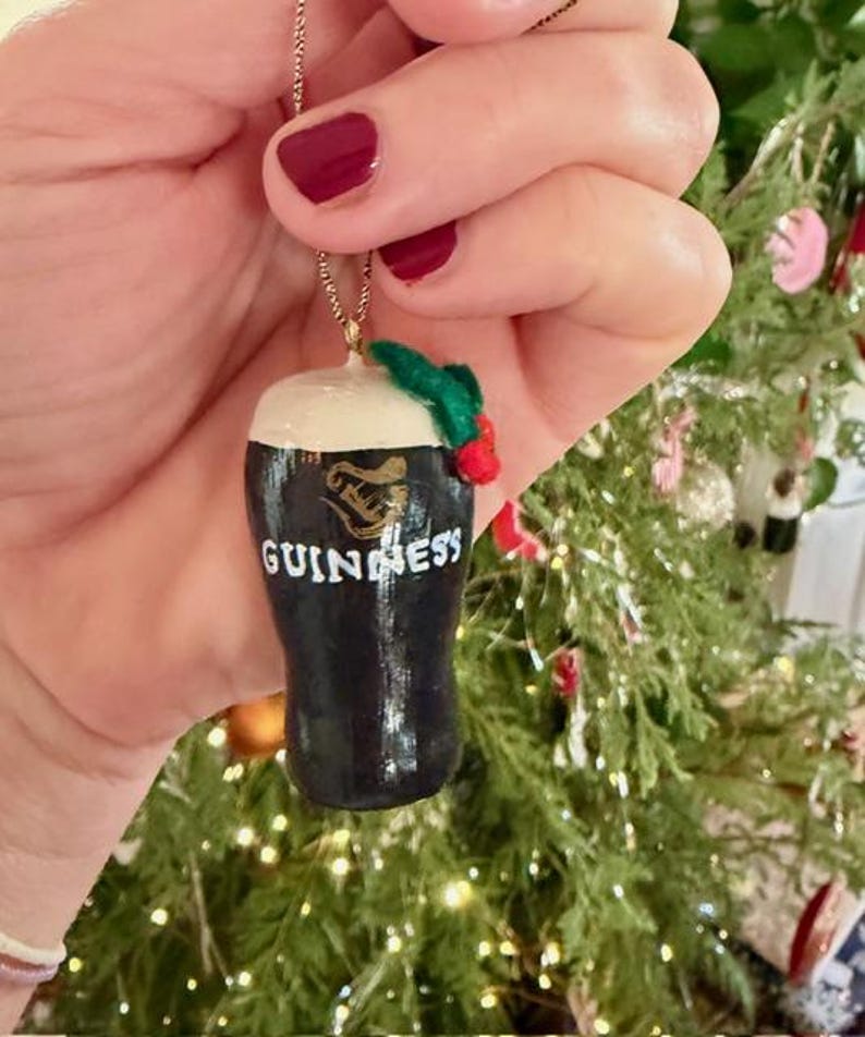 Pode incluir: Um enfeite de Natal de copo de cerveja Guinness preto e branco com uma espuma branca. O enfeite tem a palavra "Guinness" em letras brancas. Um raminho de azevinho verde e vermelho est&aacute; preso no topo.