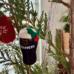 Pode incluir: Um enfeite de Natal em forma de copo de Guinness. O enfeite preto tem espuma branca e a palavra "Guinness" em branco. &Eacute; decorado com azevinho verde e bagas vermelhas, pendurado por uma corrente dourada.