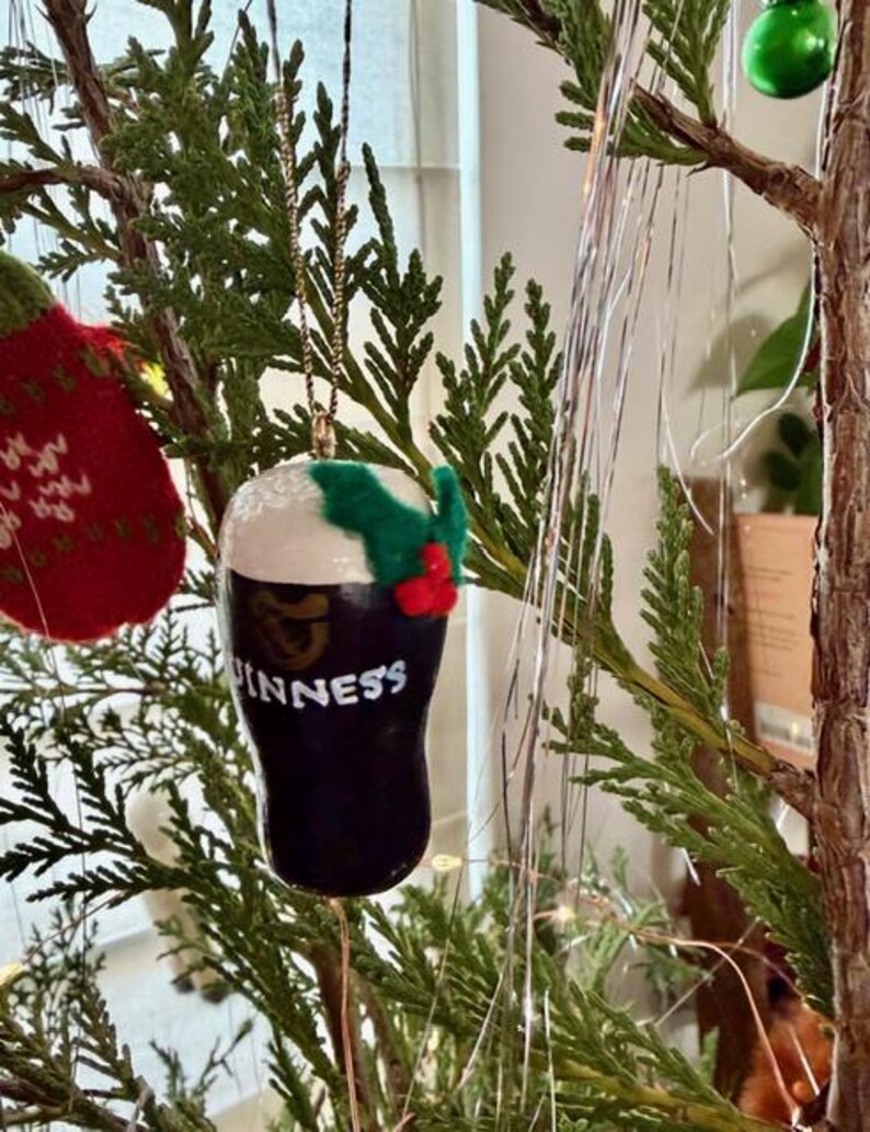 Pode incluir: Um enfeite de Natal em forma de copo de Guinness. O enfeite preto tem espuma branca e a palavra "Guinness" em branco. &Eacute; decorado com azevinho verde e bagas vermelhas, pendurado por uma corrente dourada.