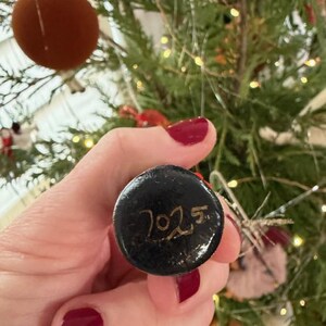 Pode incluir: Uma bola de Natal preta com o ano "2025" escrito em dourado. O enfeite redondo &eacute; segurado em uma m&atilde;o com esmalte vermelho. Uma &aacute;rvore de Natal &eacute; vis&iacute;vel ao fundo.