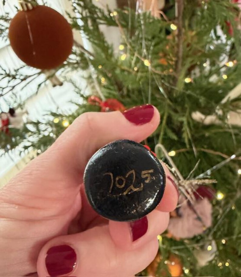 Pode incluir: Uma bola de Natal preta com o ano "2025" escrito em dourado. O enfeite redondo &eacute; segurado em uma m&atilde;o com esmalte vermelho. Uma &aacute;rvore de Natal &eacute; vis&iacute;vel ao fundo.