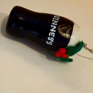 Pode incluir: Enfeite de Natal de copo de cerveja Guinness preto e branco com o logotipo da Guinness. O enfeite tem um topo de espuma branca, folhas de feltro verdes e detalhes de bagas vermelhas. Uma corda prateada est&aacute; presa para pendurar.