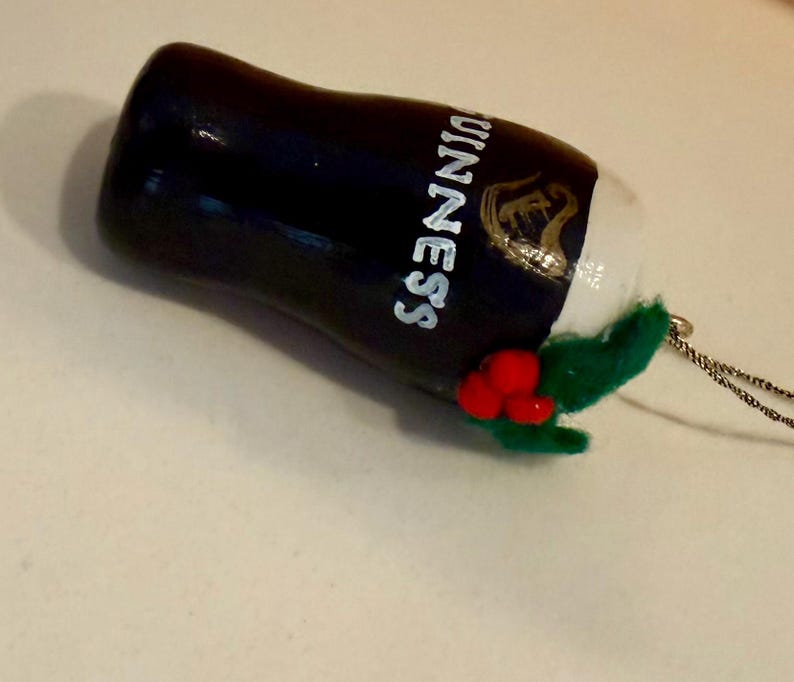 Pode incluir: Enfeite de Natal de copo de cerveja Guinness preto e branco com o logotipo da Guinness. O enfeite tem um topo de espuma branca, folhas de feltro verdes e detalhes de bagas vermelhas. Uma corda prateada est&aacute; presa para pendurar.
