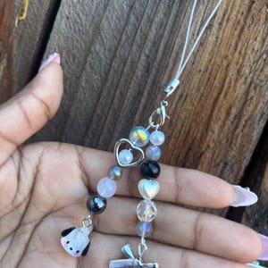 May include: A beaded phone charm with a variety of beads in shades of pink, gray, black, and iridescent. The charm includes a small dog charm, a heart-shaped bead, and a silver bow charm. The charm is attached to a white cord.